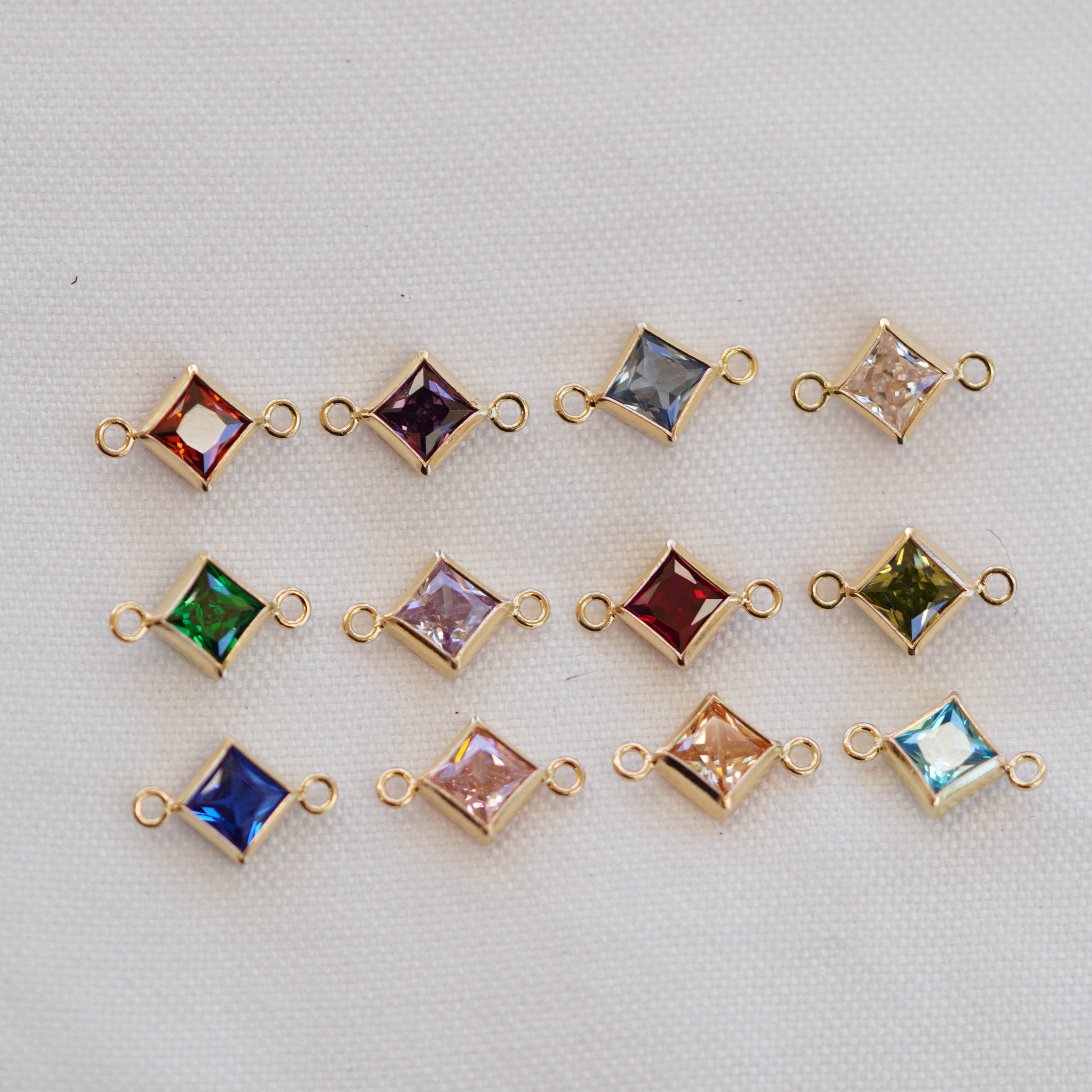 A collection of square-shaped, gold-filled birthstone connectors for jewelry making, displayed in a variety of colors corresponding to different months.