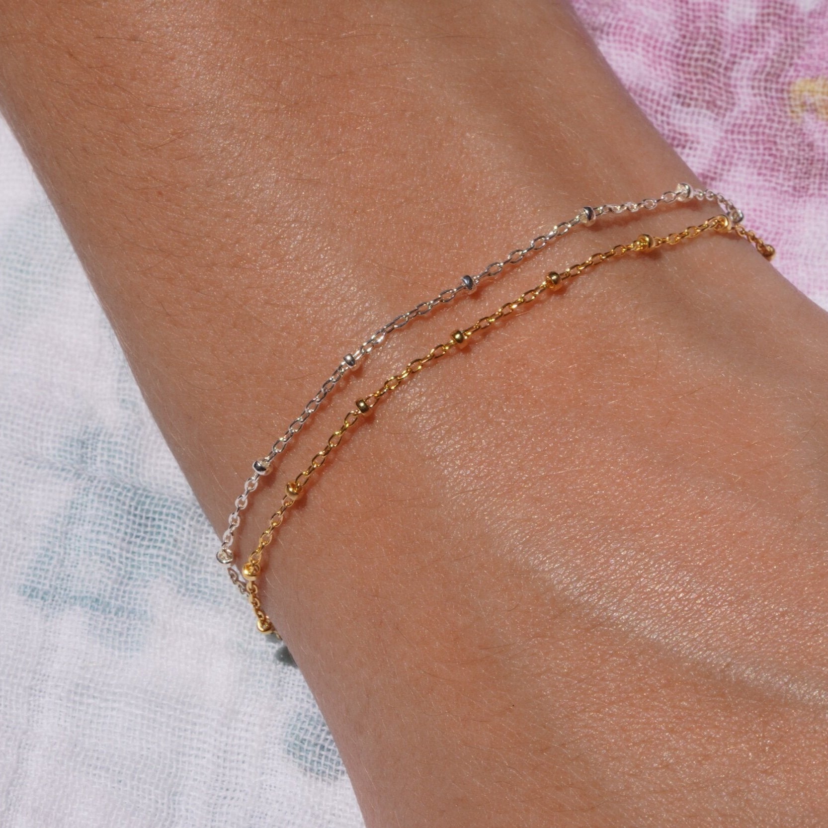 Two dainty cable satellite chains on a person's wrist, one appears to be gold filled and the other sterling silver.