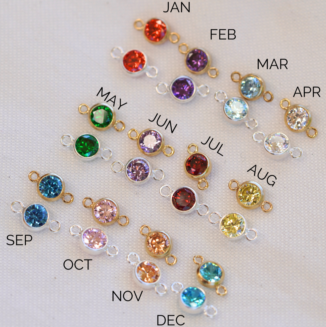 A collection of round gold filled connectors with different colored cubic zirconia stones, each labeled with a month from January to December.